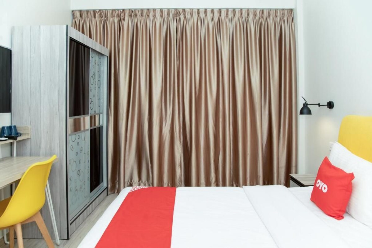 A hotel room with a bed, a desk with a chair, a window with curtains, and a bedside table with a lamp.