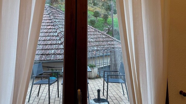 Vista attraverso una porta di vetro con tende bianche, su un patio esterno con pavimento piastrellato, un tavolo rotondo e due sedie.
