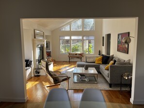 A spacious living room with a sofa, chairs, and a coffee table. There are large windows allowing natural light and a view of greenery outside.