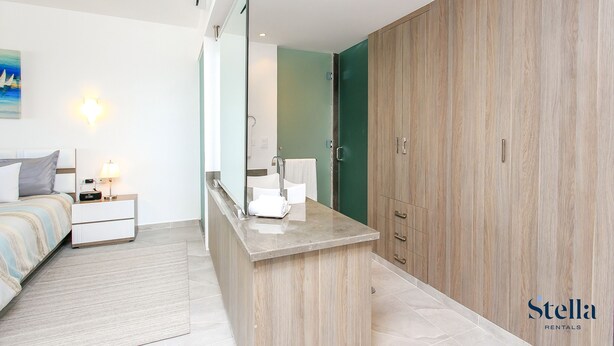 Une chambre d’hôtel moderne équipée d’un lit, d’une table de chevet, d’une commode et d’un grand miroir. On y trouve également une armoire en bois et une petite salle de bain avec un lavabo et un porte-serviettes.