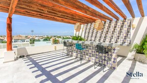 Una terraza al aire libre con una pérgola de madera, un muro con diseño y una zona de bar con dos taburetes.