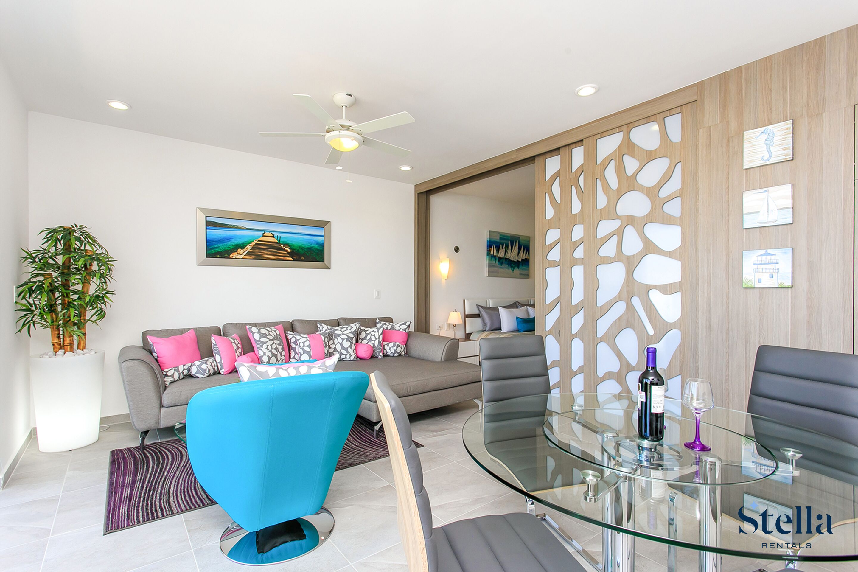 A modern living room with a sofa, a blue chair, a glass table with a bottle and glass, and a decorative wall panel.