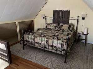 A bedroom with a sloped ceiling, a metal bed frame, a patterned bedspread, a bedside table with a lamp, and a wall-mounted shelf. - Arrow Dreams! Great location, walking distance to lake, playground & pool (Pocono Lake)