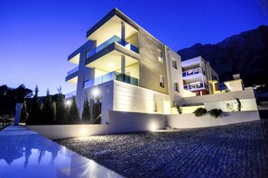 A modern multi-story building with balconies, a driveway, and a mountainous backdrop.