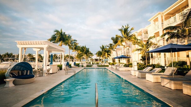 A pool area with lounge chairs and umbrellas, surrounded by palm trees and a white building. - 3 bedroom 2 bath Ocean front condo on marina with opt boat slip.   (Key West)
