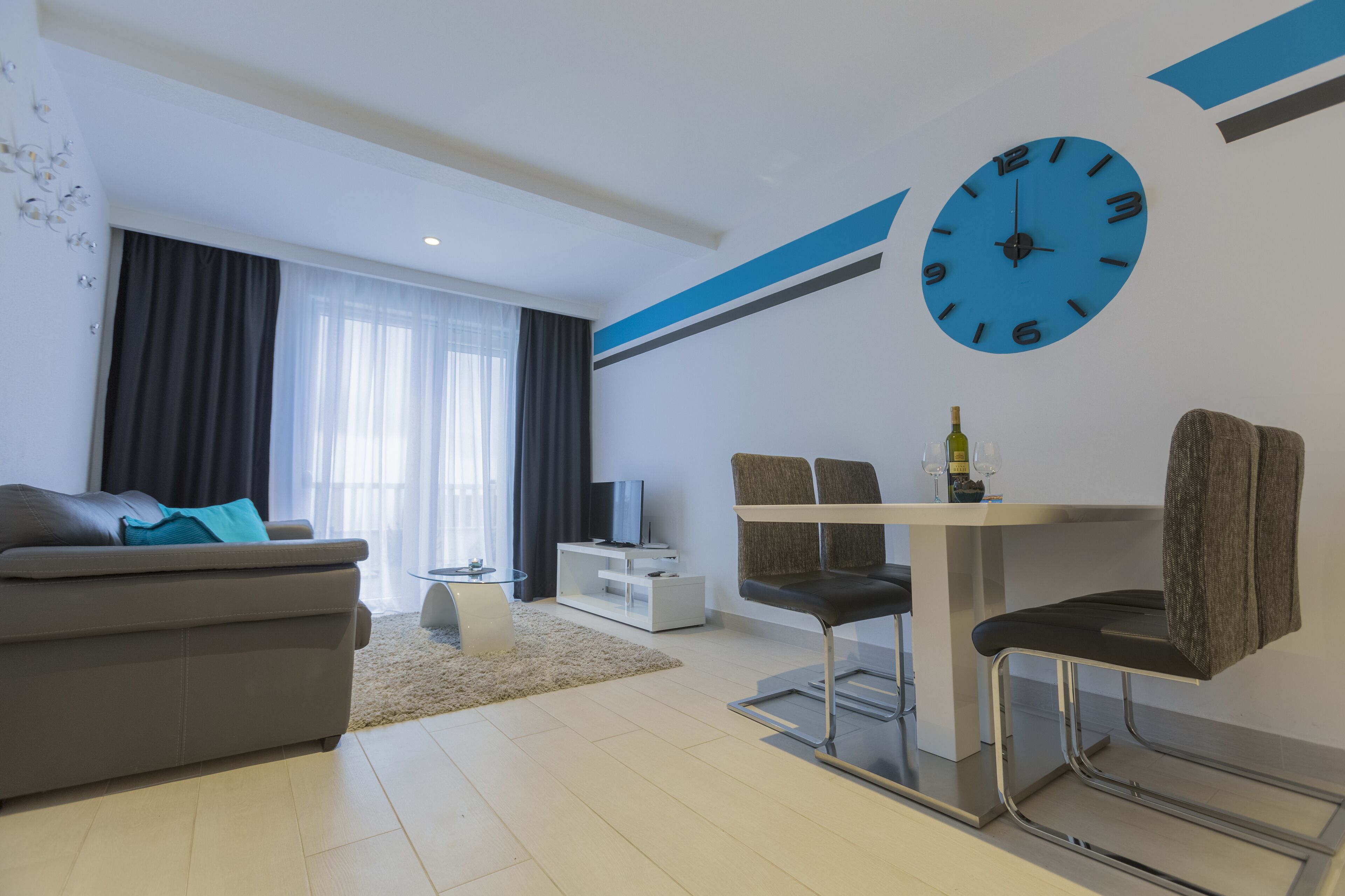 A modern living room with a sofa, a dining area with a table and chairs, a TV, and a large wall clock.