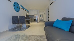 Una sala de estar moderna con un sofá gris y una almohada azul, una cocina con armarios blancos y un horno empotrado, y un reloj de pared grande.