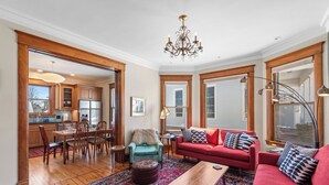 Un salon spacieux avec un canapé rouge, une table basse en bois et un tapis à motifs. On y trouve également un coin repas avec des chaises et une table en bois, et une cuisine visible à travers une porte ouverte.