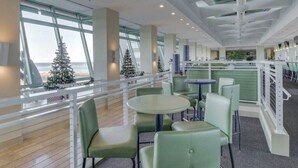 En modern inredning med runda bord och gröna stolar, stora fönster med utsikt över havet och ett vitt räcke.