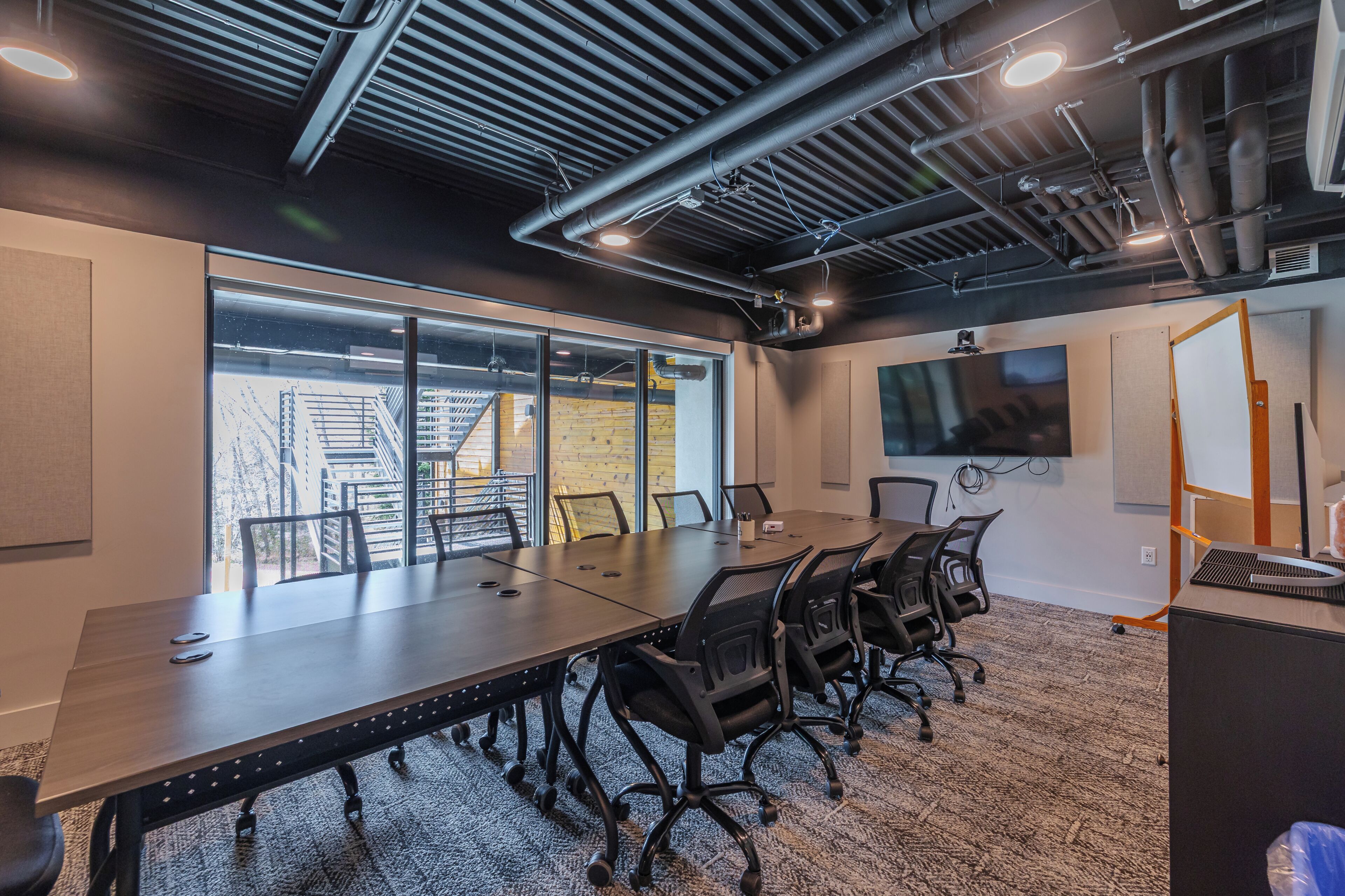 A conference room with a long table, chairs, a projector screen, and a large window with a view of outdoor stairs.