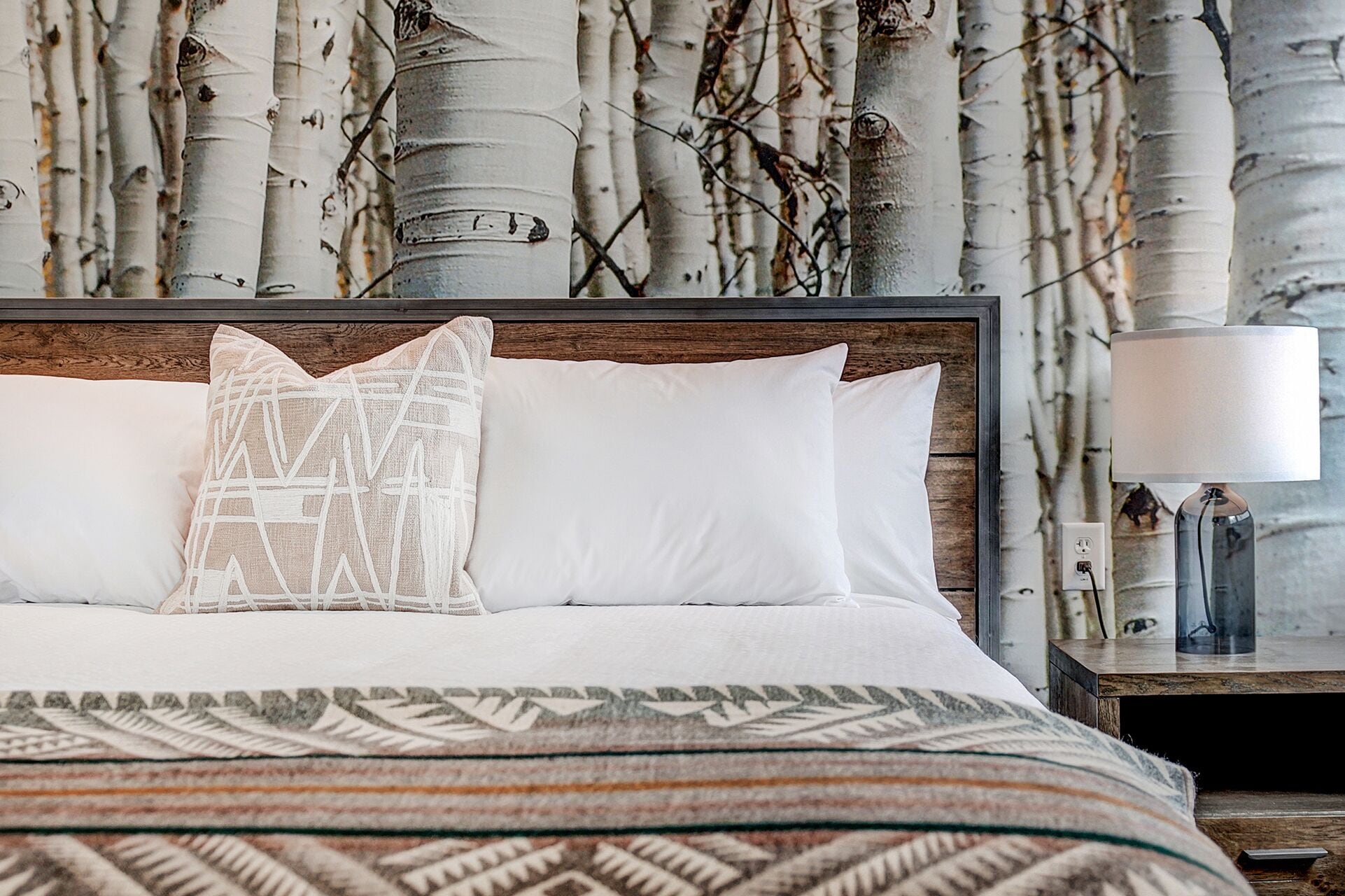 Een net opgemaakt bed met witte lakens en een gestreept plaids, een nachtkastje met een lamp, en een muur met behang van berkbomen.
