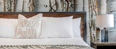 A neatly made bed with white linens and a patterned throw, a bedside table with a lamp, and a wall with birch tree wallpaper.
