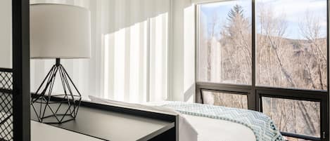 A modern bedroom with a bed, a desk, and a window with a view of trees.