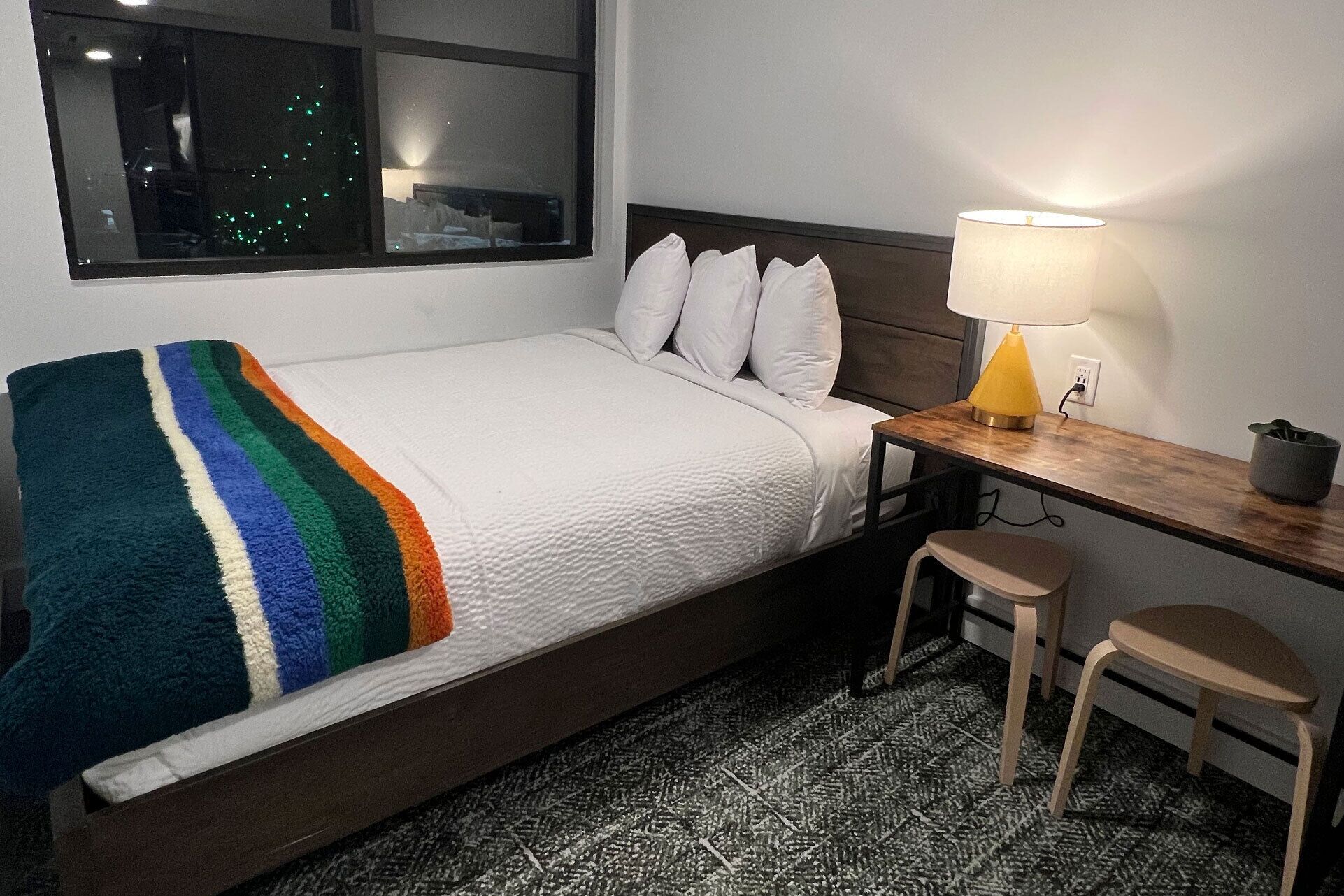 Una cama individual con edredón blanco y una manta multicolora, un escritorio de madera con una lámpara amarilla, dos sillas de madera y una ventana con vistas a un paisaje urbano nocturno.