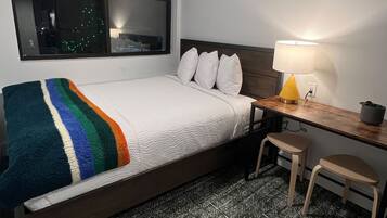 Una cama individual con edredón blanco y una manta multicolora, un escritorio de madera con una lámpara amarilla, dos sillas de madera y una ventana con vistas a un paisaje urbano nocturno.