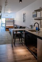 Uma cozinha moderna com uma ilha central, um espaço de refeições com bancos e uma cama beliche ao fundo.
