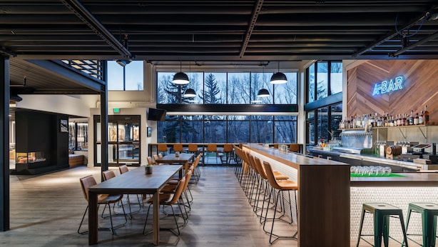 Un espace bar moderne avec des tables en bois et des tabourets de bar, de grandes fenêtres donnant sur la verdure, et un panneau néon indiquant « BAR ».