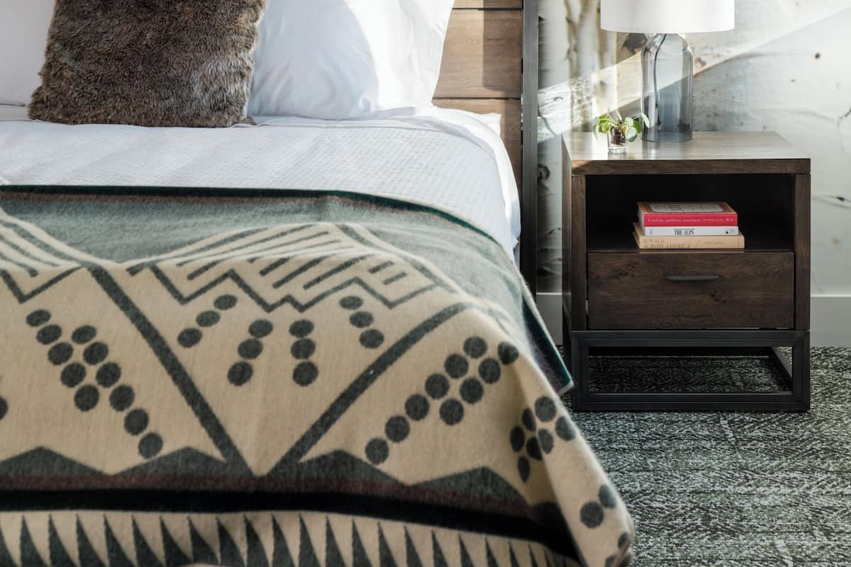 Un dormitorio con cabecera de madera, cama con ropa de cama estampada, mesita de noche con libros y pared con papel tapiz de árboles de abedul.