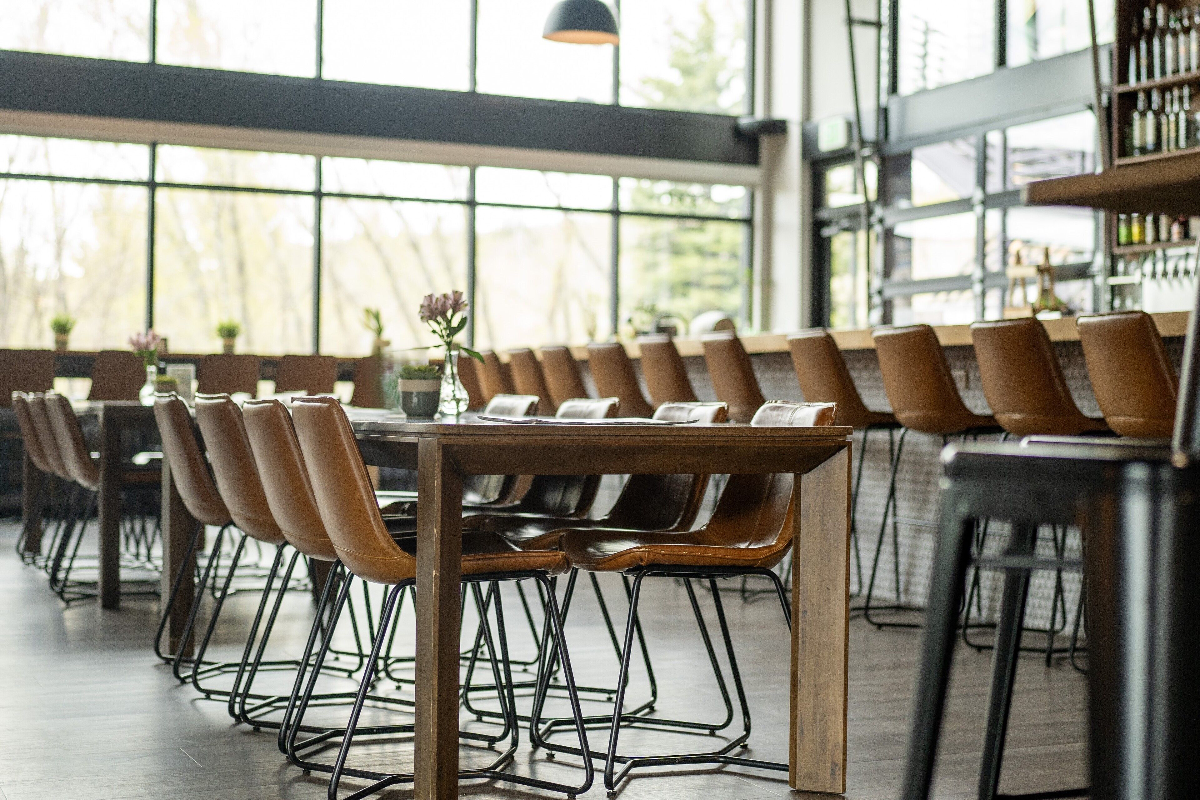 A restaurant with long wooden tables and metal chairs, large windows, and a bar area.