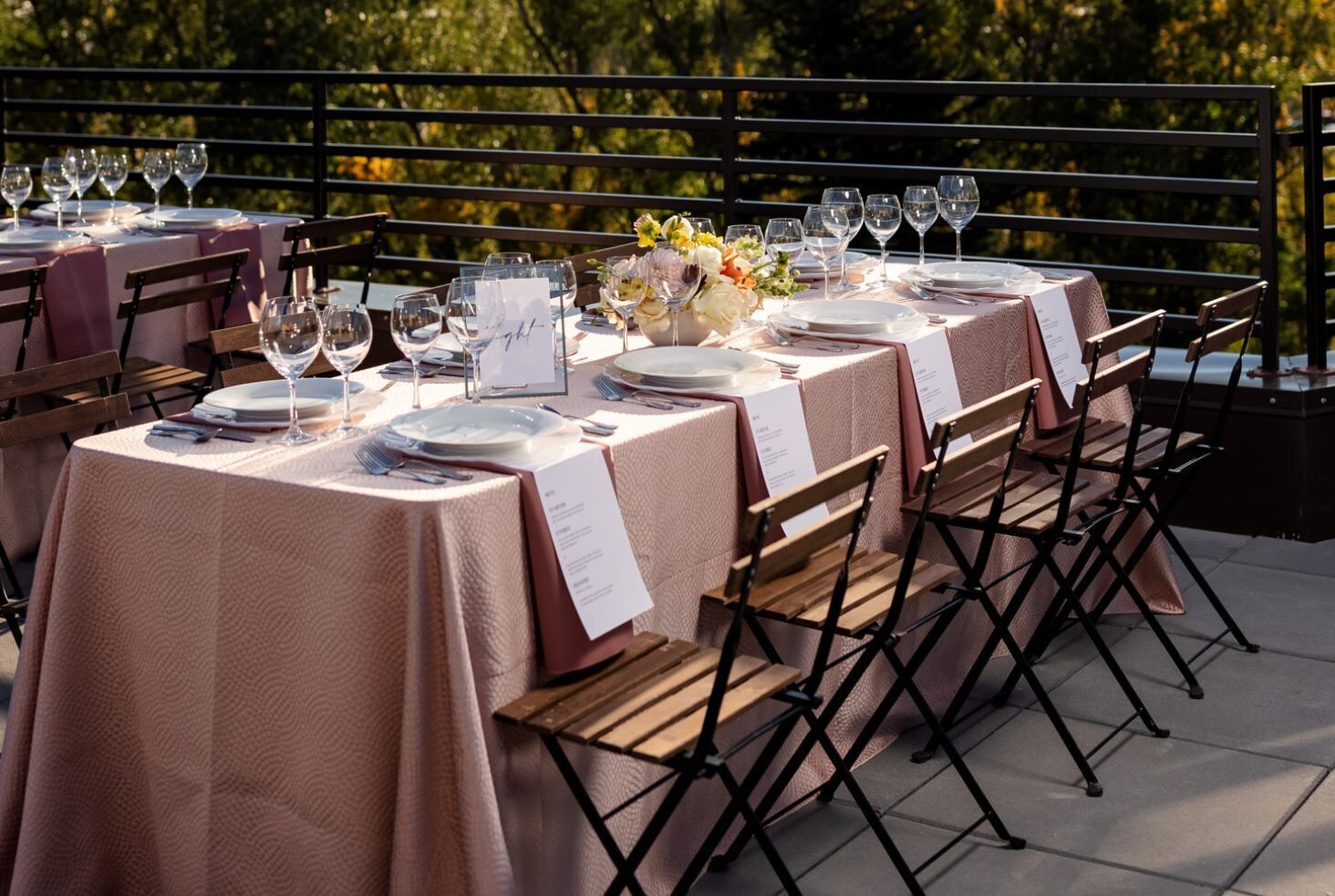 Varias mesas al aire libre dispuestas con platos blancos, cubiertos y copas de vino, cada una con una tarjeta de menú.