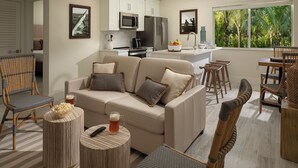 En moderne stue med en beige sofa, et spiseområde med træstole, et køkken med hvide skabe og et stort vindue med udsigt til grøn natur.