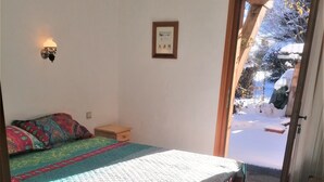 A bedroom with a bed, a bedside table, a nightstand, a wall-mounted light, a framed picture, and a view of a snowy outdoor area through an open door. - jlulbert's chalet near biotop lake and center (Combloux)