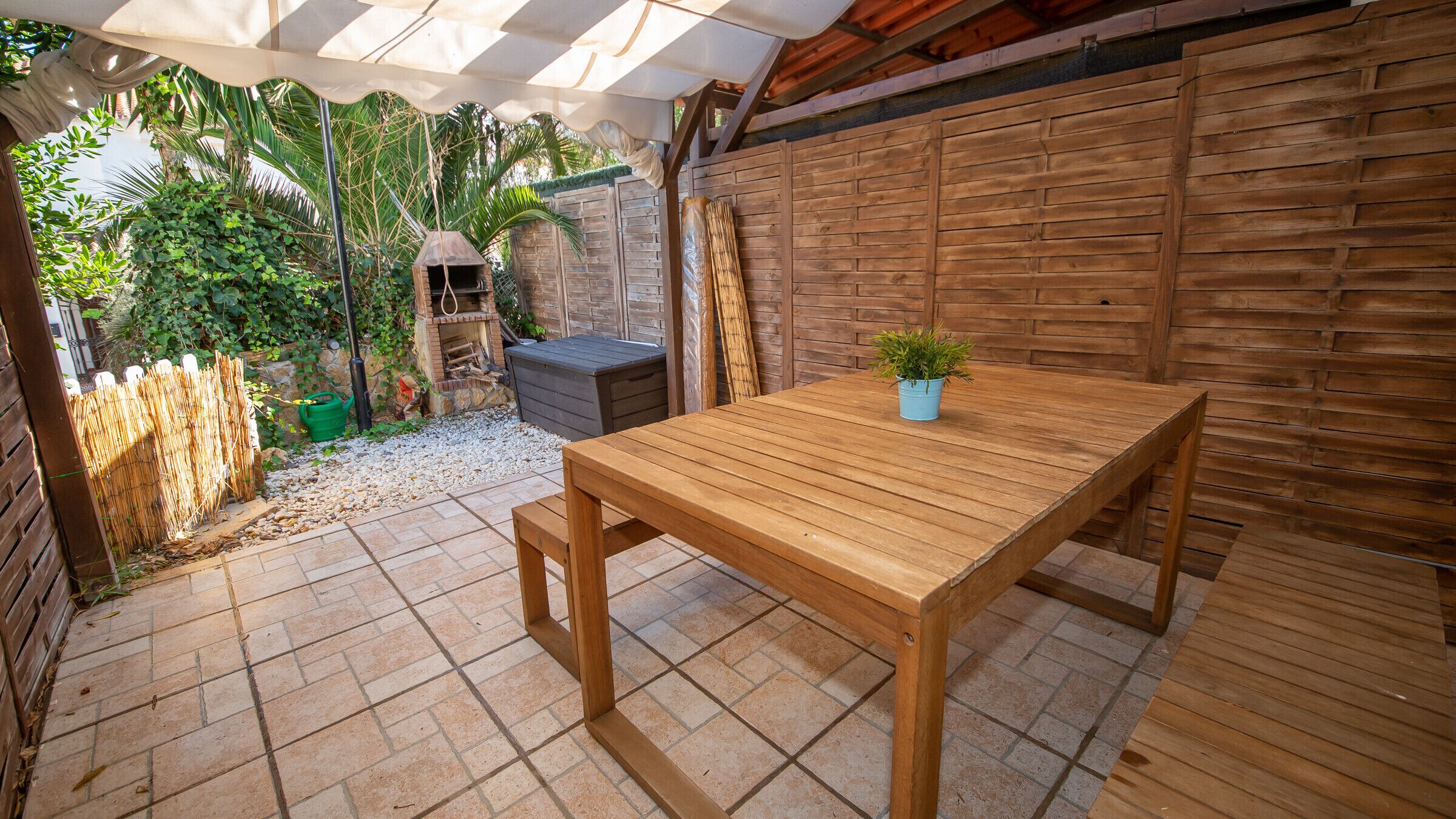 A wooden patio with a table and bench, surrounded by a bamboo fence and a stone wall.
