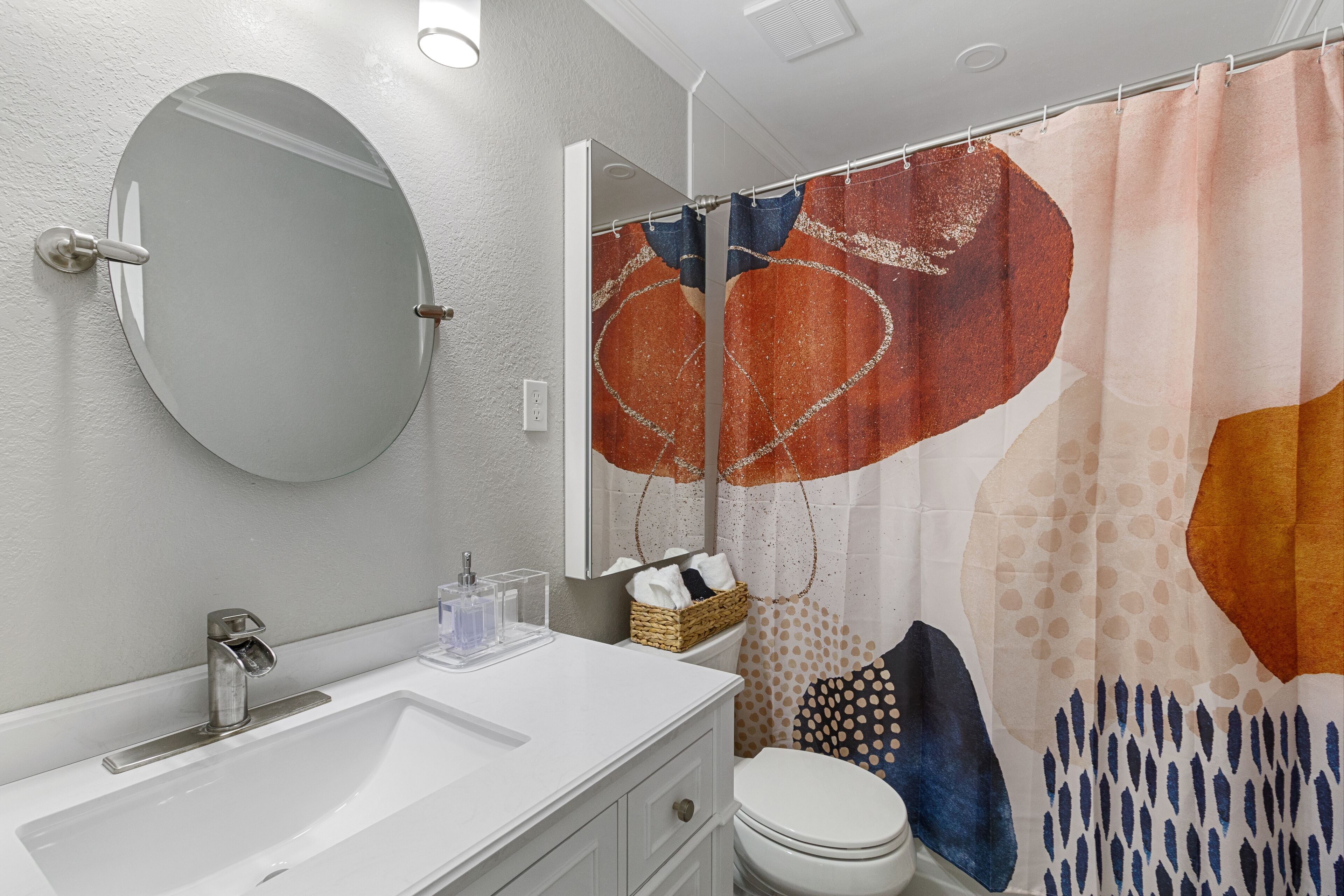 Un bagno con un lavandino bianco, uno specchio rotondo, un water e una tenda da doccia con motivi astratti.