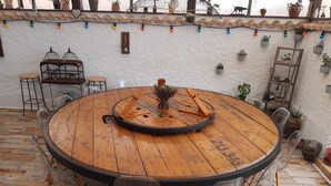 Taula rodona de fusta amb cadires, envoltada de plantes en testos i una paret decorativa.