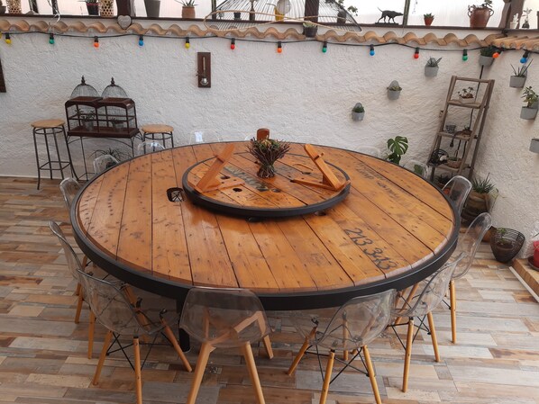 Uma mesa redonda de madeira com cadeiras, rodeada por plantas em vasos e um painel decorativo.