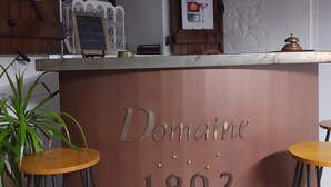 Zona de bar amb un cartell que diu "Domaine 1902" i una il·lustració d'una ampolla de vi. Hi ha dos tamborets i una planta en test.