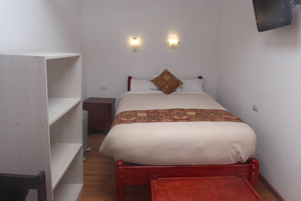 Ein kleines Zimmer mit einem Einzelbett, einem Holzbettgestell, einem Nachttisch, einem Kleiderschrank und einem an der Wand befestigten Fernseher.