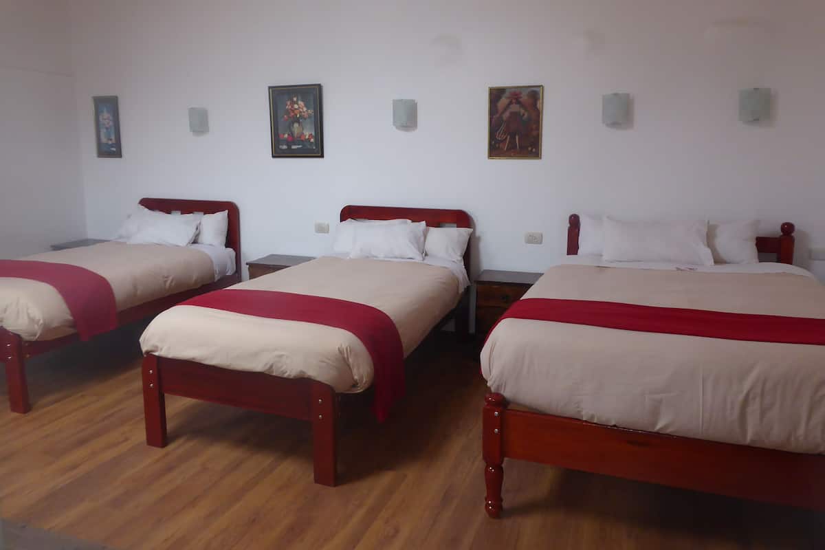 Ein Hotelzimmer mit drei hölzernen Betten, jedes mit einer roten und beigen Tagesdecke. An der Wand befinden sich Lichtschalter und gerahmte Bilder.