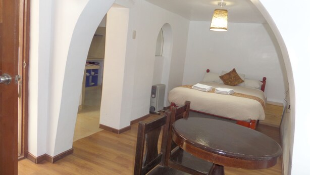 A hotel room with a wooden floor, a bed with white bedding and a brown pillow, a small table with a chair, and a ceiling light fixture. - Pirwa B&B Cusco (Cusco)
