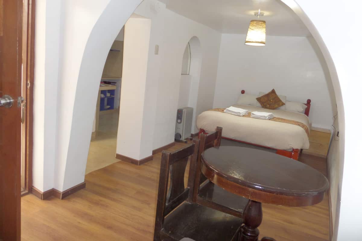 Ein Hotelzimmer mit Holzboden, einem Bett mit weißer Bettwäsche und einem braunen Kissen, einem kleinen Tisch mit Stuhl und einer Deckenleuchte.