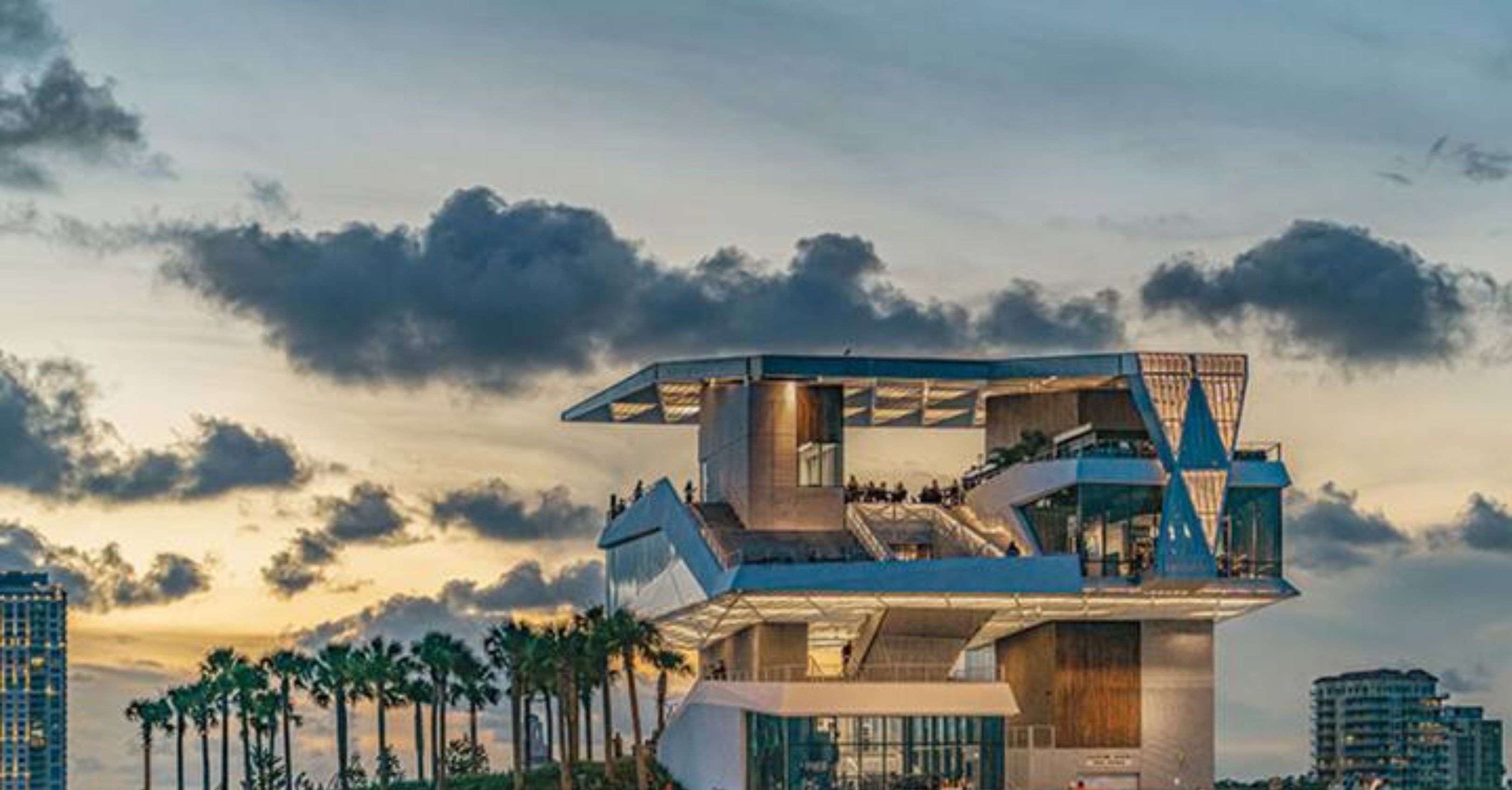 A modern waterfront hotel with a unique architectural design, featuring multiple levels and balconies, located near a pier with palm trees.