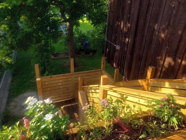 Relax on the deck surrounded by a lush garden with a large tree and a variety of plants.