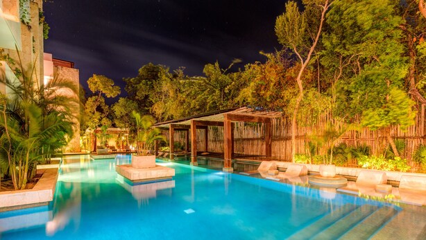 Zona de piscina nocturna con muelle de madera, sillas de descanso y valla de bambú.