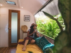 Una habitación con cama, una puerta, una mesita con un bolso tejido, y una ventana que da a zonas verdes.
