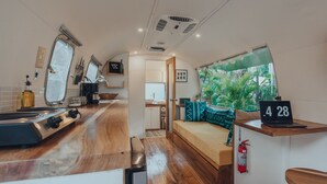 Una cocina compacta con encimera de madera, una ventana con vistas a zonas verdes y una pequeña zona de estar con una computadora portátil mostrando la hora.