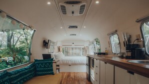 Interior de una caravana compacta con cama, zona de estar, kitchenette y ventana con vistas a la vegetación.