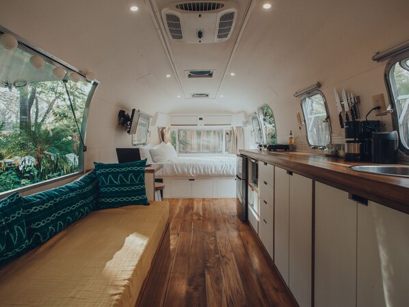 Interior de una caravana compacta con cama, zona de estar, kitchenette y ventana con vistas a la vegetación.