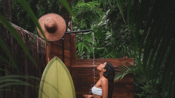 Una persona en traje de baño y sombrero de paja, de pie dentro de una estructura de madera, con una tabla de surf.