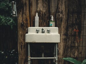 Un lavamanos blanco con dos llaves, instalado en una pared de madera con cañerías visibles y una rejilla metálica.