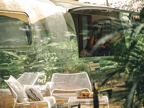 Una furgoneta camper vintage con un letrero que dice "International", sillas de mimbre blancas, una mesita con una copa de vino y una bandeja de madera con manzanas.