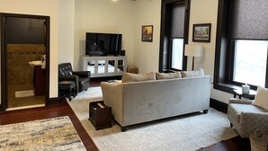 A living room with a grey sofa, a television on a cabinet, a ceiling fan, and a window with blinds. - Condo directly on Broadway!  Location, location, location!!  Walk to everything! (Saratoga Springs)