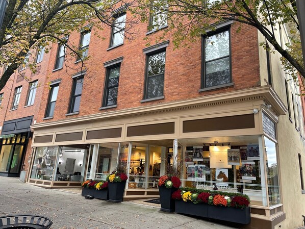 A brick building with a storefront, large windows, and outdoor seating area with flower arrangements.
