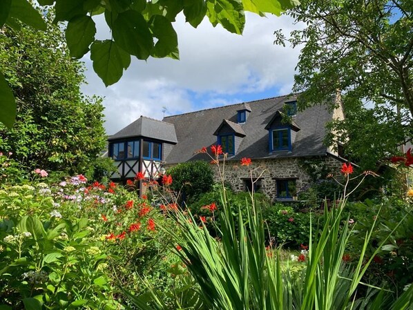 Une maison traditionnelle en pierre, avec un toit sombre, entourée d’une végétation luxuriante et de fleurs aux couleurs vives.