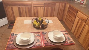 A wooden kitchen table with two white bowls and plates, a fruit basket, and a placemat with a mountain design. - Cozy cabin in a peaceful place (Ruidoso)