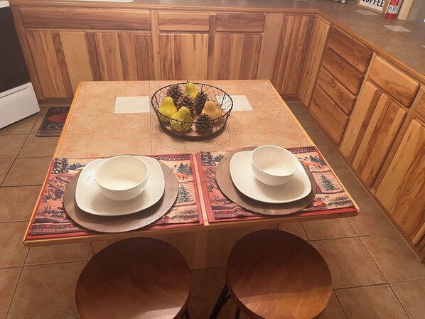 A wooden kitchen table with two white bowls and plates, a fruit basket, and a placemat with a mountain design.